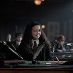 Uśmiechnięta uczennica w szacie Hogwartu, część obsady "Harry Potter i Kamień Filozoficzny", siedzi przy ławce z piórem i kałamarzem.