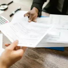 Dwie osoby analizują dokumenty, szukając informacji, gdzie zgłosić podatek od nieruchomości. Na stole kalkulator i lupa.