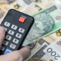 Dłoń z pilotem naciska przycisk "POWER" na tle polskich banknotów. Symbolizuje to koniec abonamentu RTV i oszczędności.