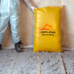 Osoba w kombinezonie stoi obok worka z materiałem do ocieplenia celulozą. Podłoga pokryta jest już warstwą izolacji.
