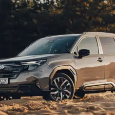 Brązowy Subaru Forester na piaszczystym podłożu, z lasem w tle.