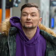 Mężczyzna w fioletowej bluzie i kurtce moro z futrzanym kapturem, jakby szukał wolności w miejskim zgiełku.