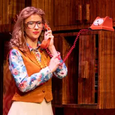 Kobieta w okularach i kwiecistej bluzce rozmawia przez czerwony telefon. Scena z spektaklu "Sylwester w Teatrze Warszawa".