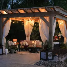 Przytulna pergola z oświetleniem i zasłonami. Zastanawiasz się, jakie drewno na pergole będzie najlepsze? Ta drewniana konstrukcja wygląda na solidną i naturalną.
