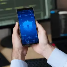 Ręce trzymające smartfon z ikoną tarczy i klucza. W tle monitory z kodem. Szukasz sposobu, jak usunąć blokadę rodzicielską w telefonie?