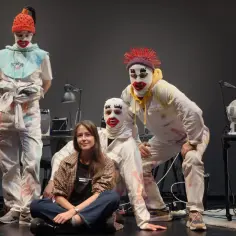 Scena teatralna z trzema postaciami w białych kombinezonach i maskach klaunów. Jedna postać siedzi, dwie stoją. To może być interludium przed głównym aktem.