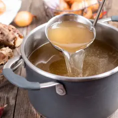Nabieranie chochlą aromatycznego, złocistego rosołu kolagenowego z garnka. Obok miska z kośćmi wołowymi i cebulą.