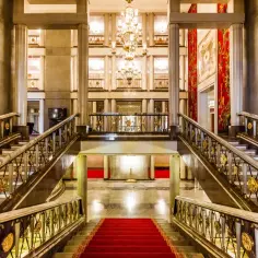 Eleganckie foyer teatru z marmurowymi schodami, złotymi balustradami i kryształowym żyrandolem. Czerwony dywan zaprasza do środka.