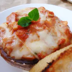 Pieczarki z grilla z mozzarellą i sosem pomidorowym, udekorowane listkiem bazylii. Obok kromka chleba.
