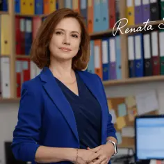 Kobieta w niebieskiej marynarce stoi przed regałem z kolorowymi segregatorami.