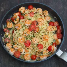 Makaron spaghetti z krewetkami, pomidorkami koktajlowymi i natką pietruszki w sosie na patelni.