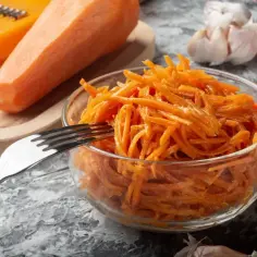 Świeża surówka z marchewki, przygotowana na tarce, czeka w szklanej misce na widelcu. Obok leżą czosnek i papryczka chili.
