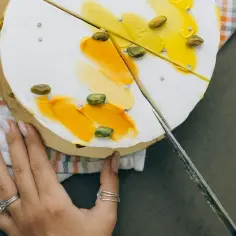 Dłoń kroi tort na 30 osób. Biały krem, żółte dekoracje i pistacje.