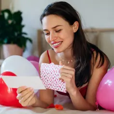 Uśmiechnięta kobieta czyta list, otoczona balonami w kształcie serc. Może to być list do męża, który rani, ale jej mina sugeruje coś innego.