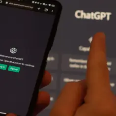 Dłoń wskazuje na ekran telefonu z logowaniem do ChatGPT. Obok widać ekran z napisem ChatGPT.