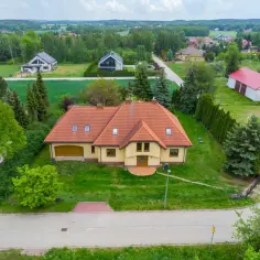 Widok z lotu ptaka na żółty dom z czerwoną dachówką, otoczony zielenią i innymi budynkami w podmiejskiej wsi.