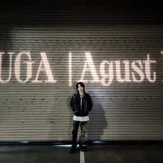 Agust D, czyli Suga z BTS, stoi przed rolowaną bramą z napisem "SUGA | Agust D" wyświetlonym na niej.