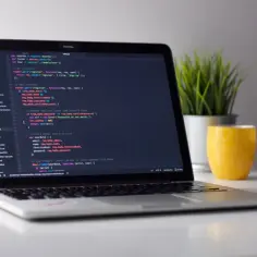 Laptop z kodem HTML na ekranie, obok zielona roślina i żółty kubek.