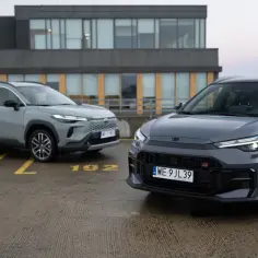 Dwie nowe Toyoty Corolla Cross, jedna szara, druga ciemnoszara GR, zaparkowane na mokrym parkingu.