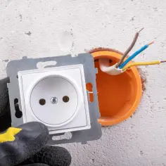 Ręka w rękawicy przygotowuje podłączenie gniazdka. Widać przewody wychodzące z puszki instalacyjnej.