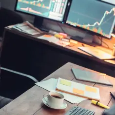 Biurko z trzema monitorami wyświetlającymi wykresy giełdowe. Kawa i notatki na blacie. Dowiedz się, jak przenieść okno na drugi monitor.