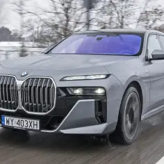 Szary BMW G70 pędzi po mokrej drodze. Elegancka sylwetka i charakterystyczny grill budzą podziw.