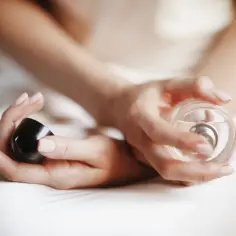 Dłonie kobiety trzymające flakonik perfum i czarną nakrętkę.