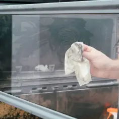 Czyszczenie szyby w kominku. Ręka z papierowym ręcznikiem usuwa sadzę z zabrudzonej szyby.