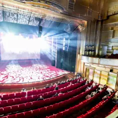Widownia Teatru Capitol wypełniona widzami, siedzącymi w czerwonych fotelach. Na scenie grają światła, tworząc magiczną atmosferę.