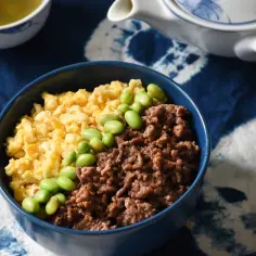 Miseczka z soboro, jajecznicą i edamame, obok czajniczka i filiżanki z herbatą, na tle tkaniny z wzorem tie-dye.