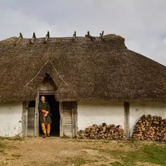Mężczyzna w stroju Anglosasów stoi przed chatą krytą strzechą.