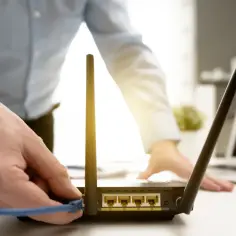 Ręka podłącza kabel do routera, przygotowując się do tego, jak zresetować router. W tle widać laptopa.