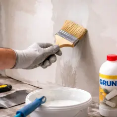 Ręka w rękawicy nakłada grunt pędzlem na tynk gipsowy. Obok stoi butelka z napisem "GRUNT" i wiaderko z zaprawą.
