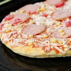 Mrożona pizza z serem, salami i kukurydzą na czarnej blasze do pieczenia.