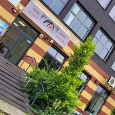 Elegancki budynek z witrynami okien i drzwi, zielenią i tablicą "OKNA ROLETy DRZWI PODŁOGI".
