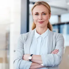 Uśmiechnięta bizneswoman w szarym żakiecie z założonymi rękami. Pokazuje, co można robić po studiach ekonomicznych: budować karierę.