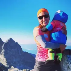 Justyna Kowalczyk z dzieckiem na szczycie górskim, uśmiechnięta, w otoczeniu skalistych szczytów i błękitnego nieba.