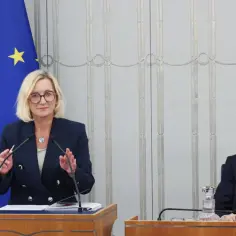 Kobieta przemawia przy mównicy obok flag Polski i UE, mężczyzna siedzi przy stole.