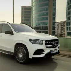 Biały Mercedes GLS 2025 pędzi po miejskiej drodze, mijając nowoczesne budynki.