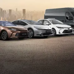 Linia modeli Toyoty: brązowa Camry Hybrid, srebrny Prius Plug-in Hybrid, biała Corolla i szary Proace Max na tle miasta.