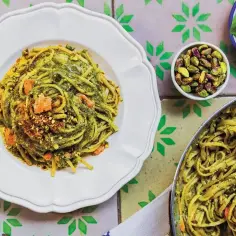Pyszne pesto z makaronem, posypane pistacjami i kawałkami łososia. Obok miseczka z pistacjami i listkami bazylii.