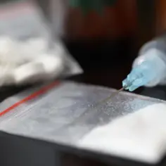 Strzykawka z igłą zanurzona w białym proszku, prawdopodobnie amfetamina, obok woreczka z tabletkami.