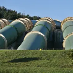 Ogromne rury elektrowni szczytowo pompowej, zielone i beżowe, wznoszą się na tle lasu i błękitnego nieba.