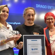 Trzy osoby odbierają nagrodę. Kobieta po lewej trzyma szklanego byka, a mężczyzna w środku, z logo DRAGO ENTERTAINMENT na koszulce, prezentuje dyplom.
