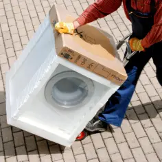 Pracownik w kombinezonie i rękawicach transportuje zapakowaną pralkę na wózku. Na kartonie widnieje napis "MAX 600 kg", co sugeruje, ile waży pralka.