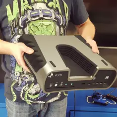 Mężczyzna trzyma konsolę PS5, porównując ją z możliwościami PC. Na niebieskiej szafce leży kontroler i pilot.