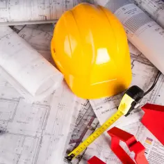 Plany budowlane, żółty kask, miarka, poziomica i czerwone modele domów.