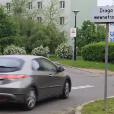 Szary samochód jedzie po drodze wewnętrznej obok zieleni i budynków.