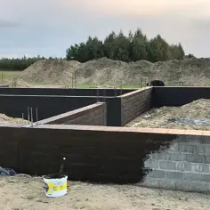 Fundament domu w budowie, częściowo pokryty ciemną izolacją, z widocznym wiadrem farby.