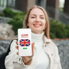 Uśmiechnięta dziewczyna trzyma telefon z napisem "LEARN ENGLISH" i flagą Wielkiej Brytanii. To darmowa aplikacja do nauki angielskiego.